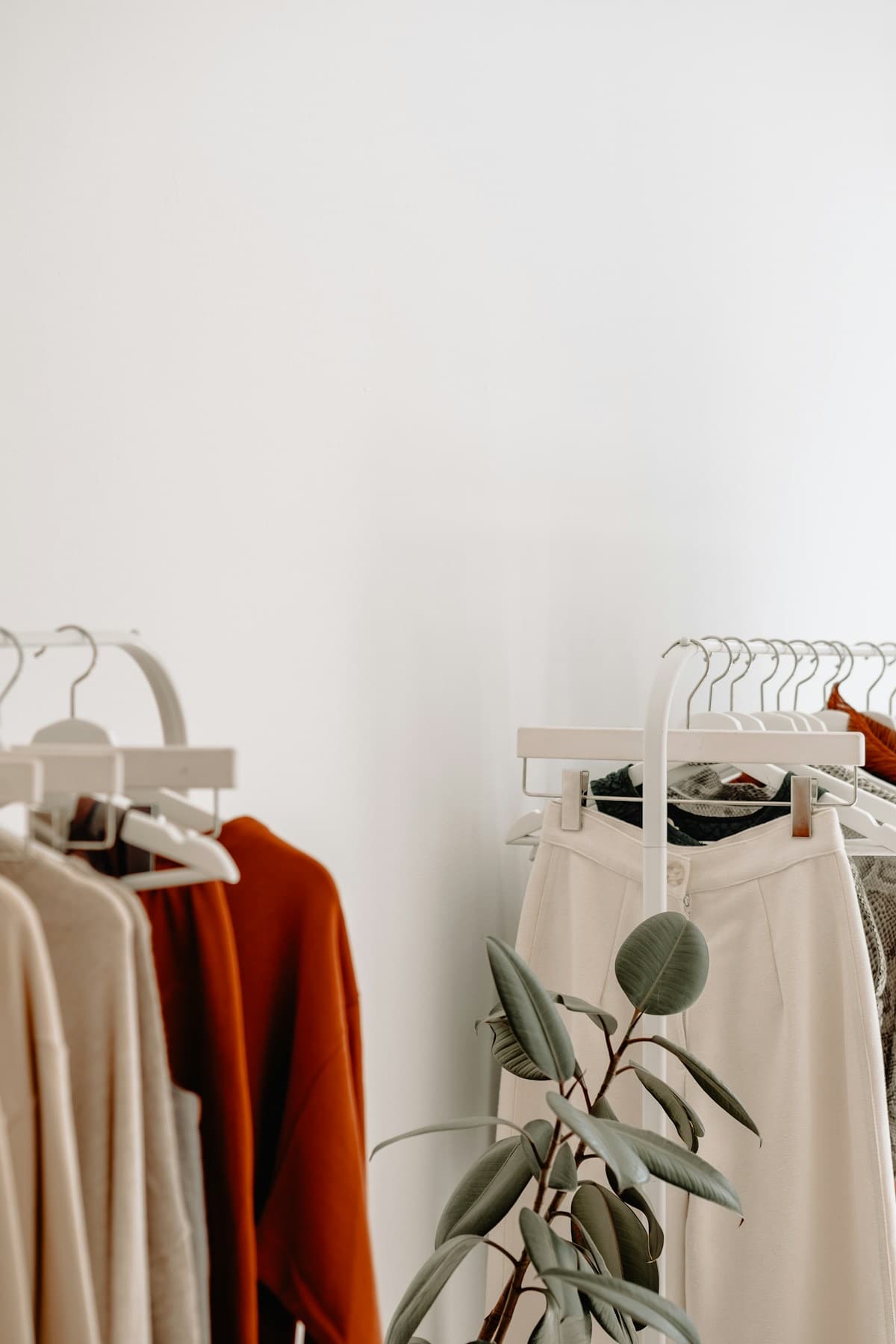 Two white clothing racks holding neutral and rust-colored garments beside a potted plant.